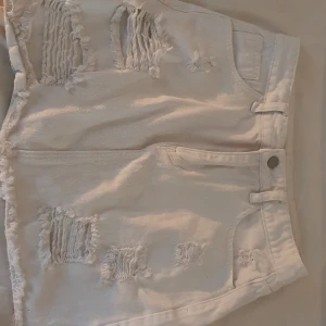 Vit slitna jeanskjol  - Snygga vita jeansshorts med slitna detaljer och fransig kant. Klassisk femficksmodell med bälteshällor och knappstängning. Perfekta för en chill sommarlook och passar till allt. Råa slitningar framtill ger en trendig vibe.