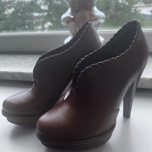 Bruna klackade boots från UGG, stl 36 - Snygga bruna boots från UGG i skinn med hög klack och plattform. Insidan är fodrad med mjuk ull för extra komfort. Dekorativ flätad kant runt öppningen ger en unik touch. Perfekta för dig som vill ha både stil och värme.