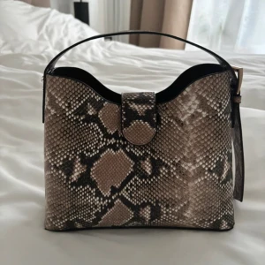 Axelväska i ormmönstrat skinn - Snygg axelväska med ormmönster i beige, brunt och svart. Väskan har ett brett handtag, guldfärgade detaljer och dragkedja på insidan. Perfekt för dig som vill sticka ut med en trendig accessoar. Rymmer allt du behöver till vardagen.