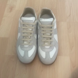 Maison Margiela vita sneakers - Maison Margiela sneakers i vitt skinn med ljusgrå mockadetaljer och beige snörning. Snygg gummisula i brunt ger en klassisk look. Perfekta för dig som gillar stilrena och trendiga skor med premiumkänsla.