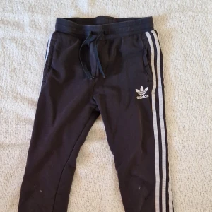 Svarta Adidas mjukisbyxor - Svarta mjukisbyxor från Adidas med klassiska vita ränder längs sidorna och Adidas-logga på låret. Byxorna har snörning i midjan..