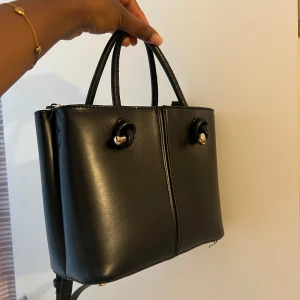 Svart handväska med gulddetaljer och band  - Stilren svart handväska i skinnimitation med två handtag och justerbar axelrem. Pris kan diskuteras vid snabbköp 