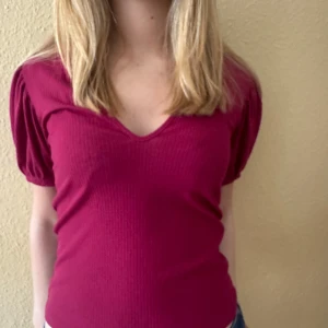 Mörkrosa ribbad topp med puffärm - Snygg mörkrosa topp med v-ringning och korta puffärmar. Ribbad struktur ger en cool och trendig känsla. Perfekt att matcha med jeans eller kjol för en avslappnad men stilsäker look.