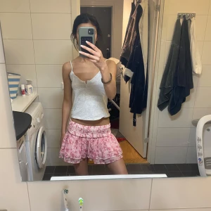 Rosa volangkjol med mönster - Supersöt kort volangkjol i rosa med blommigt mönster och bred smock i midjan. Kjolen har flera lager och ger en fluffig, lekfull look. Perfekt för varma dagar och enkel att styla med en enkel topp.