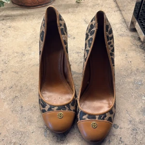 Leopardmönstrade pumps från Tory Burch - Snygga pumps från Tory Burch med leopardmönstrad textil och bruna skinn-detaljer. Rund tå med guldfärgad logga framtill. Klack och insida i brunt skinn, perfekt för dig som vill sticka ut med en cool och trendig look.