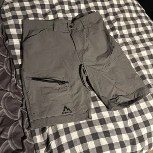 Grå McKinley shorts med dragkedja - Snygga grå shorts från McKinley i syntetmaterial, perfekta för sommarens äventyr. De har en praktisk ficka med dragkedja på benet och klassisk passform med bälteshällor. Stilrena och funktionella för dig som gillar att vara aktiv utomhus.