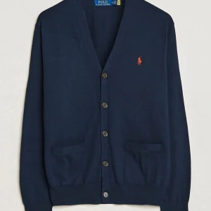 Marinblå kofta från Polo Ralph Lauren - Klassisk marinblå kofta från Polo Ralph Lauren i bomull med v-ringning, knappar framtill och två fickor. Ikoniska röda loggan broderad på bröstet. Perfekt för lager-på-lager och snygg till både jeans och chinos.