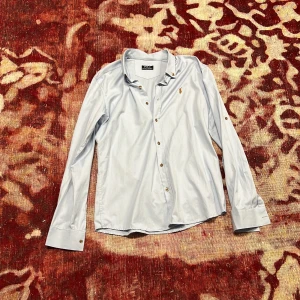 Ljusblå skjorta från Polo Ralph Lauren - Klassisk ljusblå skjorta från Polo Ralph Lauren med broderad logga på bröstet. Skjortan har lång ärm, knappar framtill och på kragen samt stilren passform. Perfekt för dig som gillar en clean och tidlös look.