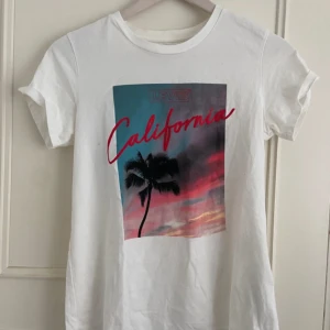 KÖP FRÅN MIN VINTED - Vit t-shirt från Levi's med färgglatt tryck av en palm och texten 'California' i rött. Klassisk rund hals och korta ärmar. Perfekt för en chill och avslappnad stil med sommarkänsla.