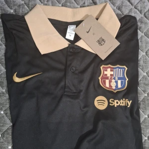 Svart FC Barcelona fotbollströja Nike - Snygg svart FC Barcelona fotbollströja från Nike med guldfärgade detaljer och klubbmärke på bröstet. Tröjan har en beige krage och knappar framtill samt Spotify-logga. Perfekt för dig som vill visa ditt stöd för Barça.