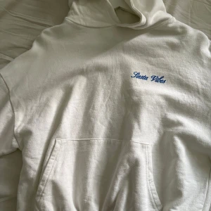 Vit hoodie från Pull&Bear med tryck - Vit hoodie från Pull&Bear med blått och gult tryck på ryggen och liten blå text på bröstet. Stor känguruficka framtill och huva.