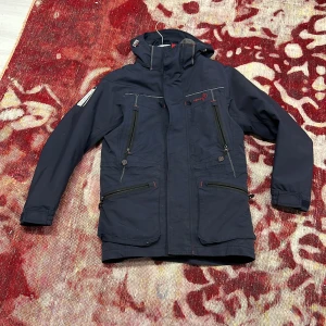 Marinblå vindjacka från Pelle P XS - Snygg marinblå vindjacka från Pelle P med huva, flera dragkedjeförsedda fickor och coola röda detaljer. Jackan har lång ärm, hög krage och praktiska justeringar vid ärmslut. Perfekt för dig som gillar funktionell och stilren design.