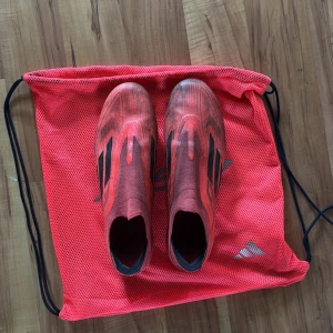 Röda adidas fotbollsskor utan snörning - Säljer ett par röda adidas fotbollsskor med svarta detaljer och klassiska tre ränder på sidan. Skorna har en strömlinjeformad design utan snörning och är tillverkade i syntetmaterial för bästa grepp och känsla på planen. Perfekta för dig som vill sticka ut på matchen.