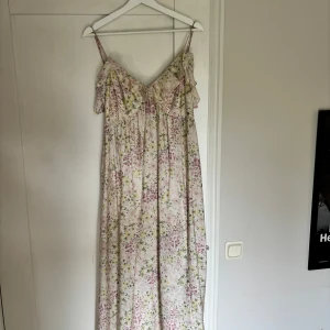 Blommig långklänning från H&M - Romantisk långklänning från H&M med tunna axelband och volangdetaljer vid bysten. Klänningen har ett ljust blommigt mönster i rosa, gult och grönt på vit botten. Perfekt för sommarens alla tillfällen och ger en riktigt dreamy vibe.