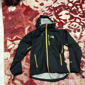 Svart vindjacka The North Face Summit - Svart vindjacka från The North Face Summit Series med gula dragkedjor och detaljer. Jackan har huva, flera fickor med dragkedja och justerbara muddar. Perfekt för dig som gillar outdoor och vill ha en snygg och funktionell jacka.