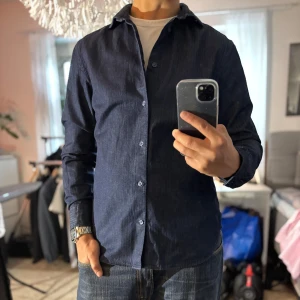 Mörkblå slim fit skjorta från Riley - Snygg mörkblå skjorta från Riley i slim fit-modell. Skjortan har diskreta ränder, klassisk krage och knäppning framtill. Långärmad och tillverkad i ett mjukt bomullstyg som känns bekvämt hela dagen. Perfekt för dig som gillar stilrena och moderna plagg.