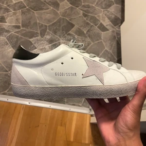 Golden Goose vita sneakers med stjärna - Snygga vita sneakers från Golden Goose med ikonisk grå stjärna på sidan och svart detalj vid hälen. Skorna har grå snörning och en slitstark sula med fiskbensmönster. Tillverkade i skinn och mocka för en lyxig känsla. Perfekta för dig som gillar streetstyle.. dust bag tillkommer