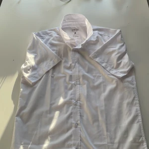 Vit kortärmad skjorta från SMOG - Klassisk vit skjorta från SMOG i regular fit med korta ärmar och knappar framtill. Skjortan är tillverkad i ett lättskött material och har en stilren, enkel design som passar perfekt till jeans eller chinos.