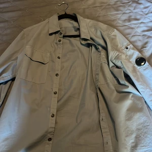 Overshirt från C.P. Company - Snygg  overshirt från C.P. Company med klassisk krage, bröstficka och ikonisk linsdetalj på ärmen. Skjortan har knäppning framtill och är tillverkad i ett lätt material som passar perfekt för lager-på-lager-stil.