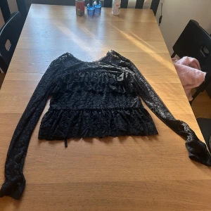 Svart spetsblus med volanger - Supersnygg svart blus i spets med långa ärmar och flera volanglager framtill. Transparent och detaljerad med blommigt spetsmönster, perfekt för dig som gillar en romantisk och edgy stil. Rund halsringning och figurnära passform.