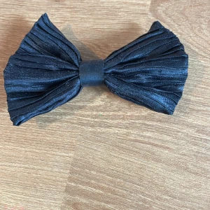 Svart plisserat hårspänne med rosett - Snyggt svart hårspänne med stor plisserad rosett i tyg. Fästs enkelt med metallspänne på baksidan. Perfekt accessoar för att lyfta din look med en trendig touch. Rosetten har en cool struktur och ger en elegant vibe.