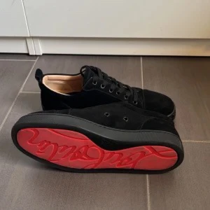 Loubs - Stilrena svarta sneakers i mocka med klassisk snörning och en unik röd sula med mönstrad text. Skorna har rund tå och platt sula, perfekt för dig som vill sticka ut med en cool detalj under foten.