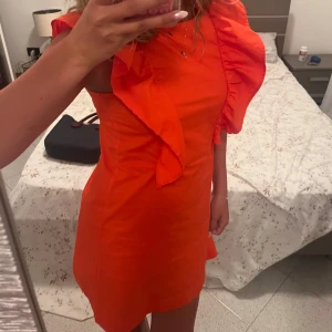 Orange kortklänning med volang - Säljer denna jättefina klänning som jag älskar men som tyvärr börjat bli lite liten. Den har volanger och är lite öppen i ryggen med en söt rosett.  Den funkar på mig som vanligtvis har xs/s men skulle snarare att säga att den är en xxs.