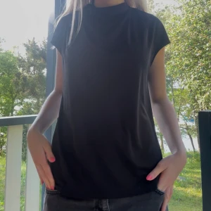 Flowy svart blus 🖤 - En svart blus med lös passform och hög, draperad hals. Toppen har korta ärmar och en snygg detalj med knapp och liten öppning i nacken. 