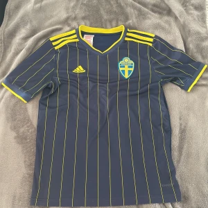 Svensk landslagströja Adidas blå/gul - Säljer en blå fotbollströja från Adidas med gula detaljer och tunna gula ränder. Tröjan har Sveriges landslagsemblem på bröstet och klassiska Adidas-ränder på axlarna. Kortärmad modell i lätt och ventilerande material, perfekt för match eller träning.