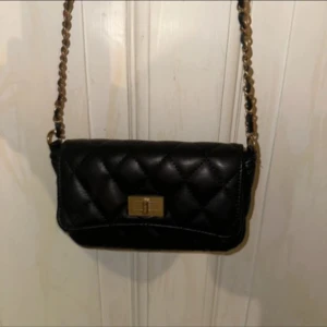 Svart quiltad axelväska från Glitter - Säljer en svart quiltad axelväska från Glitter med guldfärgad kedjerem flätad med svart material. Väskan har ett klassiskt rutmönster, snyggt spänne framtill och kompakt format – perfekt för mobilen och det viktigaste. Stilren och trendig accessoar till din outfit.