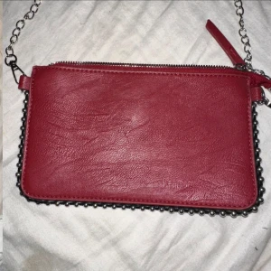 Röd axelväska med kedja - Snygg röd axelväska i skinnimitation med silverfärgad kedja som axelrem. Väskan har dragkedja upptill och coola metallkulor längs kanterna för extra edge. Perfekt storlek för det viktigaste och passar till många outfits. Alldrig använd. 