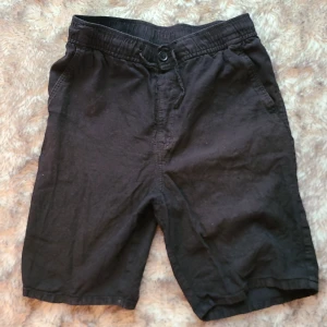 Svarta linneshorts från H&M - Svarta shorts från H&M i storlek 158. De har elastisk midja, knapp och dragkedja framtill samt fickor på sidorna och en bakficka. Materialet är en skön mix av 52% linne och 48% bomull, perfekt för varma dagar.
