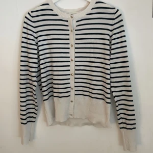 Randig kofta från Holly & Whyte - Snygg kofta i vitt med svarta horisontella ränder från Holly & Whyte. Långärmad modell med rund halsringning och knappar framtill. Tillverkad i mjuk bomull, perfekt för lager-på-lager och en klassisk look.