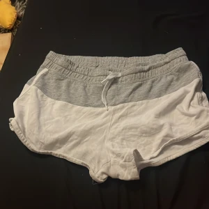 Gråvita shorts från H&M med snörning - Snygga och bekväma shorts från H&M i tvåfärgad design med grå midja och vita ben. De har elastisk midja med snörning och är gjorda i mjuk bomull, perfekta för chill dagar eller träning. Sportig look och sköna att bära hela sommaren.