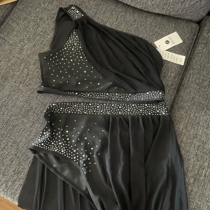 Dans dräckt  - Snygg svart långklänning från Freelee med oneshoulder-design och glittriga strassdetaljer över byst och midja. Klänningen har ett elegant fall och markerad midja, perfekt för dig som vill sticka ut med stilren glamour.