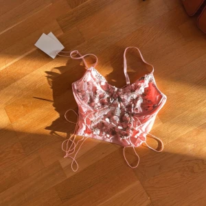 Nya Rosa blommig korsett med snörning - Superfin rosa korsett-topp med broderade blommor och tunna axelband. Toppen har transparent mesh med 3D-blommor och snörningsdetaljer på sidorna för extra snygg passform. Perfekt för dig som vill sticka ut med en romantisk och trendig look.