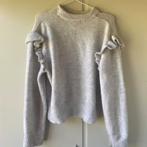 Ljusgrå stickad tröja med volang - Superfin ljusgrå stickad tröja med volangdetaljer på axlarna och längs ärmarna. Tröjan har en hög, ribbad halsringning och långa ärmar. Perfekt för dig som gillar en söt och trendig look med det lilla extra.