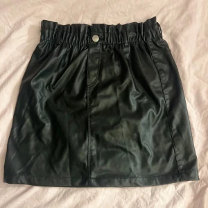 Svart kort kjol i skinnimitation - Snygg svart kort kjol i skinnimitation med resår i midjan och knapp framtill. Kjolen har en rak passform och är perfekt för att skapa en edgy look. Passar till både sneakers och boots.