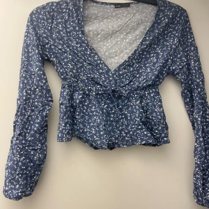 Blommig blå omlottblus med knyt - Supersöt blå blus från Bershka med vitt blommönster, omlottmodell och knyt i midjan. Djup v-ringning och långa ärmar ger en romantisk vibe. Perfekt för dig som gillar en feminin och trendig stil.