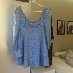 Blommig blå kortklänning med volang - Supersöt blå kortklänning med små rosa och vita blommor, volang vid halsringningen och långa puffärmar med breda manschetter. Perfekt för vår och sommar, ger en romantisk vibe och är riktigt bekväm att bära. Har prislap kvar.