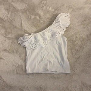 Vit offshoulder topp från Zara - Superfin vit offshoulder topp från Zara med bred volang och broderade spetsdetaljer. Toppen har en axel och är perfekt för dig som gillar romantisk och somrig stil. Mjuk bomullskänsla och snygg passform som sticker ut.