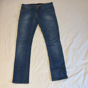 Blå jeans från Nudie Jeans, W30 L34 - Säljer ett par klassiska blå jeans från Nudie Jeans med snygga slitningar och kontrastsömmar. Modellen har raka ben och fem fickor, samt Nudie Jeans signaturbroderi på bakfickorna. Perfekta för dig som gillar en tidlös och avslappnad look.