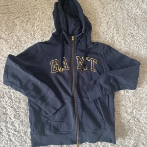 Marinblå hoodie från GANT med dragkedja - Säljer en marinblå hoodie från GANT med broderad logga i gult på bröstet. Tröjan har huva med snörning, hel dragkedja i guld och två fickor framtill. Storlek XS