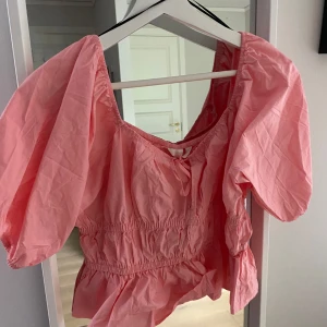 Rosa blus med puffärmar och volang - Supersöt rosa blus med puffiga korta ärmar och volang nertill. Smockad midja ger en snygg passform och den har en liten rosett framtill. Perfekt för dig som gillar romantisk och trendig stil. Liten i storleken