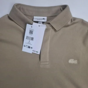 Beige pikétröja från Lacoste - Snygg beige pikétröja från Lacoste i klassisk passform. Tröjan har krage, kort knappslå och den ikoniska broderade krokodilloggan på bröstet. Tillverkad i mjuk bomull med lite elastan för extra komfort. Perfekt för en clean och stilren look. Bredd 52 cm, Längden 68cm.