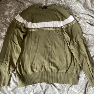 Olivgrön tröja med vit rand - Olivgrön tröja från H&M med en bred vit rand och smala gröna linjer över bröstet. Tröjan har rund halsringning, långa ärmar och ribbade muddar vid ärmslut och nederkant. Perfekt för dig som gillar en avslappnad och stilren look.