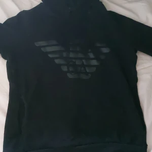 Svart hoodie från Emporio Armani - Svart hoodie från Emporio Armani med stort, diskret logotryck på bröstet. Klassisk modell med huva och dragsko. Mjuk insida och långärmad passform, perfekt för en clean och stilren look. Knappt använd. 4 gånger. 