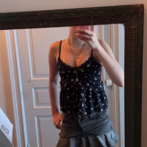 Svart blommig med spets - Supersöt sydd svart linnetopp med vita små blommor och tunna axelband. Den har en rosett för att knyta ihop tröjan där bak. Toppen har v-ringning med spetskant, markerad midja och volangig nederdel. Perfekt för dig som gillar romantisk och feminin stil. Kjolen är också till salu! 