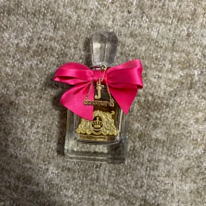 Juicy Couture Viva La Juicy parfym 100ml - Juicy Couture Viva La Juicy parfym i en lyxig glasflaska med guldiga detaljer och en stor, klar kristallformad kork. Flaskan är dekorerad med en rosa rosett och guldfärgade hängen. Innehåller 100 ml Eau de Parfum. 
