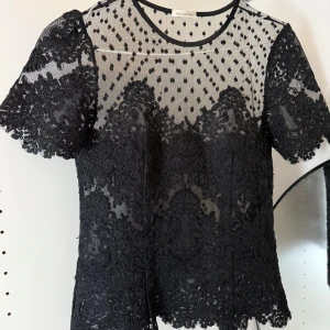 Svart spetsblus från Ida Sjöstedt - Supersnygg svart blus från Ida Sjöstedt med transparent mesh upptill och vackra spetsdetaljer över hela plagget. Korta ärmar och rund halsringning. Perfekt för dig som vill sticka ut med en elegant och trendig look.